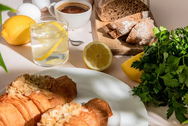 Cruasanes frescos, pan, perejil, café y agua de limón para el desayuno en la mesa gris, enfoque selectivo - foto de stock
