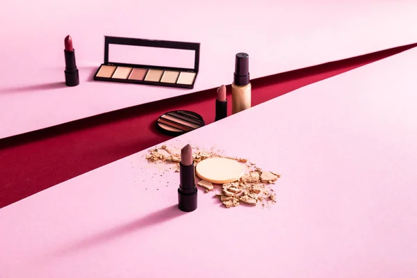 Eye shadow palettes, lipsticks and cracked face powder near face foundation on pink and crimson — Stock Photo