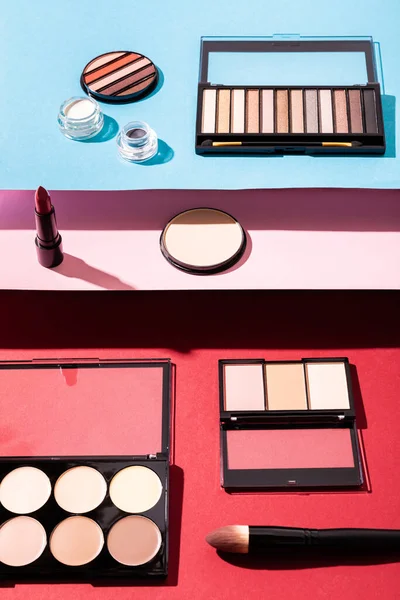 Blush and eye shadow palettes near cosmetic brushes, lipstick and face powder on crimson, pink and blue — Stock Photo