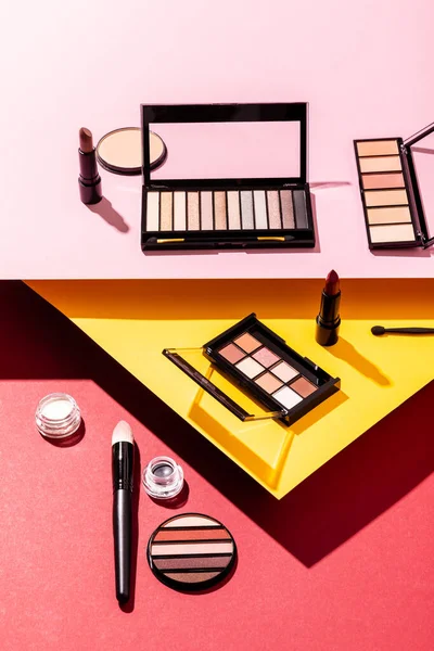 Eye shadow palettes near cosmetic brushes, lipsticks and face powder on crimson, pink and yellow — Stock Photo