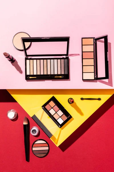 Top view of eye shadow palettes near cosmetic brushes, lipsticks and face powder on crimson, pink and yellow — Stock Photo