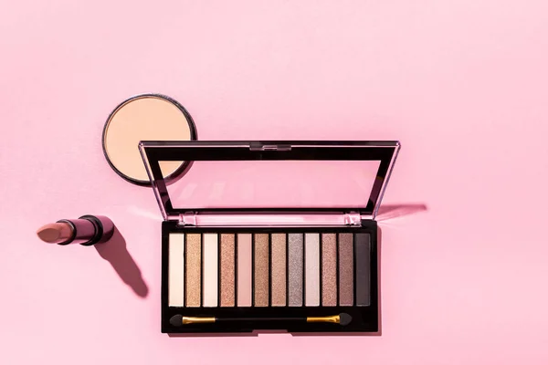 Vista superior da paleta de sombra e escova cosmética perto de batom e pó de rosto em rosa — Fotografia de Stock