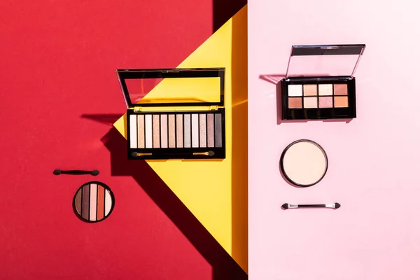 Top view of eye shadow palettes and double-sided eyeshadow applicators near face powder on crimson, pink and yellow — Stock Photo