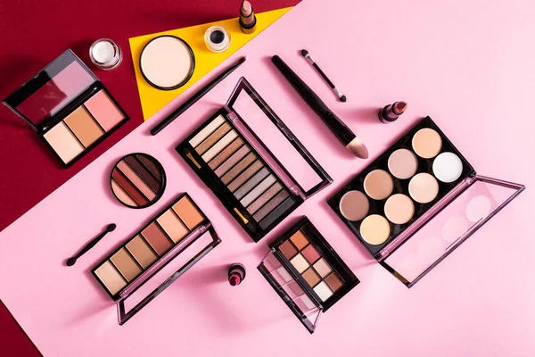 Top view of blush and eye shadow palettes near cosmetic brushes and lipsticks on pink, yellow and crimson — Stock Photo