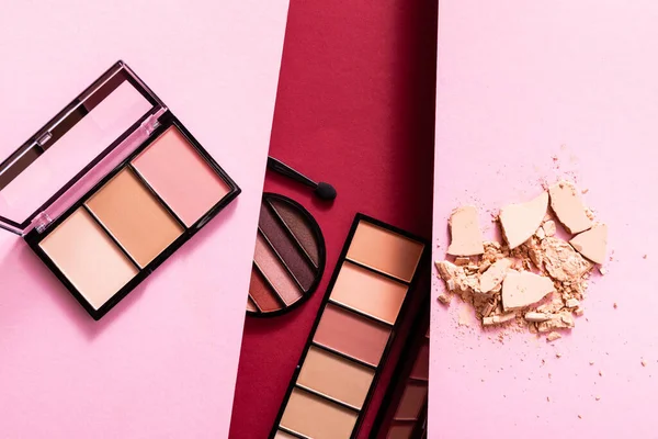 Top view of pastel eye shadow and blush palettes near cracked face powder on pink and crimson — Stock Photo