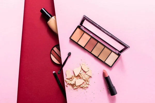 Top view of face foundation near eye shadow palette, cracked face powder, cosmetic brushes and lipstick on pink and crimson — Stock Photo