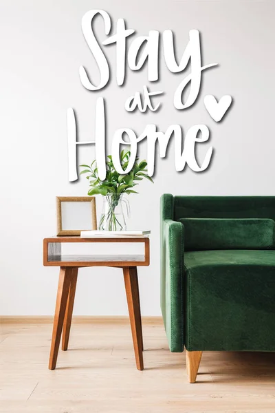 Green sofa near coffee table with plant, books, photo frame and stay at home lettering — Stock Photo