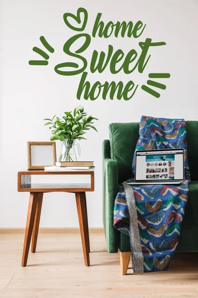 KYIV, UCRANIA - 14 DE ABRIL DE 2020: hogar dulce hogar letras cerca de sofá verde, manta, ordenador portátil con sitio web amazon y mesa de centro de madera - foto de stock