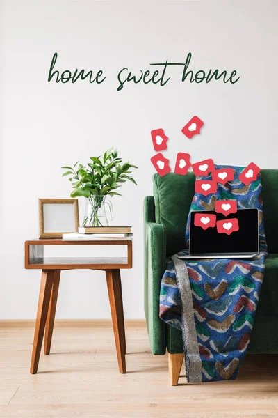 Sofá verde, manta, ordenador portátil con pantalla en blanco y corazones ilustración cerca de mesa de centro de madera con planta verde, libros, marco de fotos y hogar dulce hogar letras - foto de stock