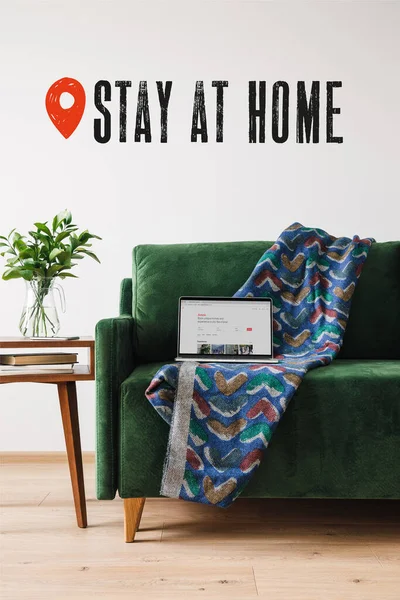 KYIV, UKRAINE - APRIL 14, 2020: green sofa, blanket and laptop with airbnb website near wooden coffee table with green plant and stay at home lettering — Stock Photo