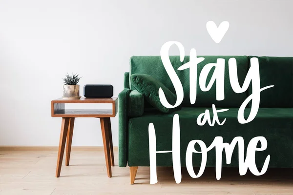 Sofá verde, almohada, mesa de centro de madera con planta y despertador cerca de quedarse en casa con letras - foto de stock