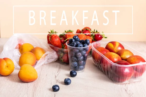 Blueberries, strawberries, nectarines and peaches in plastic containers on wooden surface near breakfast lettering on beige — Stock Photo