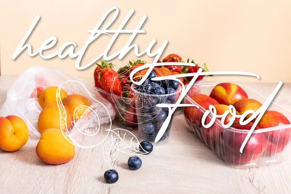 Arándanos, fresas, nectarinas y melocotones en recipientes de plástico en superficie de madera cerca de letras de alimentos saludables e ilustración de aguacate en beige - foto de stock