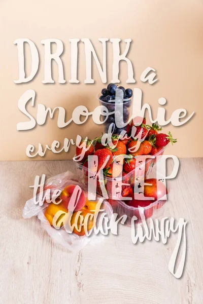 Fruit composition with blueberries, strawberries, nectarines and peaches in plastic containers near drink a smoothie every day keep the doctor away lettering on beige — Stock Photo