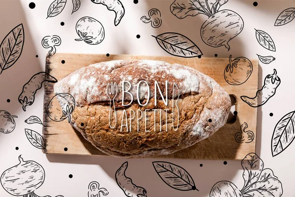 Top view of bread on cutting board on grey table with bon appetit lettering and vegetables drawing — Stock Photo