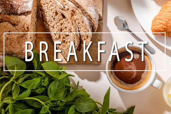 Top view of bread, basil and cup of coffee on grey table with breakfast lettering — Stock Photo