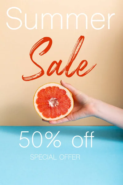 Vista recortada de la mujer que sostiene la mitad jugosa de pomelo cerca de la venta de verano, cincuenta por ciento de descuento, letras oferta especial en beige y azul - foto de stock