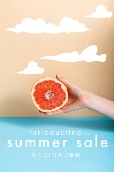 Cropped view of woman holding juicy half of grapefruit near summer sale, in stores and online lettering on beige and blue — Stock Photo