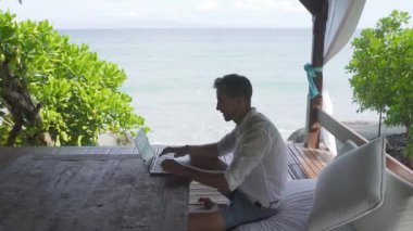 Çardak içinde oturan ve deniz manzaralı, statik kare, orta görünümü ile bilgisayarda çalışan mutlu adam