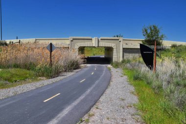 Jordan River Parkway Trail, Legacy Parkway Trail sınır Redwood Trailhead, kayalık dağlar boyunca çevredeki ağaçlar ve silt dolu çamurlu su ile panorama manzarası, Salt Lake City, Utah.