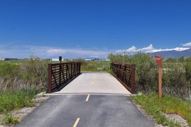 Jordan River Parkway Trail, Legacy Parkway Trail sınır Redwood Trailhead, kayalık dağlar boyunca çevredeki ağaçlar ve silt dolu çamurlu su ile panorama manzarası, Salt Lake City, Utah.