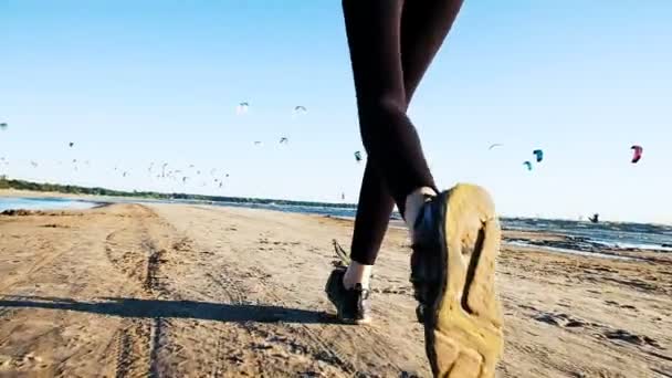 Frauenbeine in Turnschuhen in sommerlicher Großaufnahme vor der Kulisse von Kitesurfern. Frau läuft bei Sonnenuntergang im Meer, Zeitlupe. — Stockvideo
