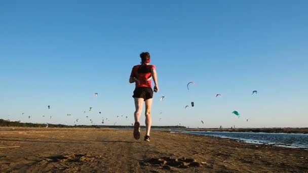 Corrida de treinamento ao pôr do sol, atleta homem corre ao longo da praia de areia contra o pano de fundo de kitesurfers, câmera lenta — Vídeo de Stock