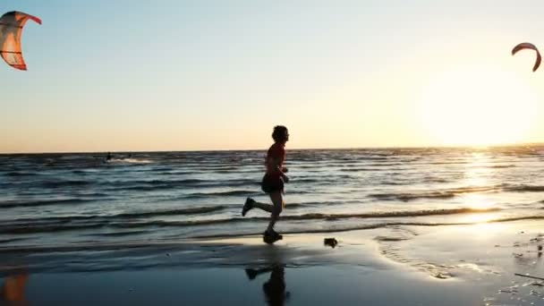 Mann läuft am Strand entlang und freut sich im Ziel vor dem Hintergrund der Kitesurfer bei Sonnenuntergang. Sportler spritzt Wasser — Stockvideo
