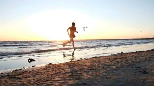 Člověk je cvičit, běžící na pláži pozadí kitesurfers při západu slunce, pomalý pohyb. — Stock video