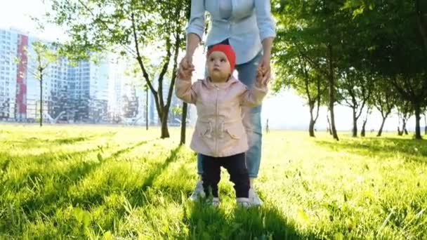 La madre ayuda a su hijo a caminar sobre la hierba en el parque al atardecer. Feliz bebé da los primeros pasos, cámara lenta . — Vídeos de Stock