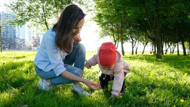 Anne ve çocuk parkta çim üzerinde günbatımında oturuyor. Mutlu aile ağır çekim bir çiçek ile oynamak.