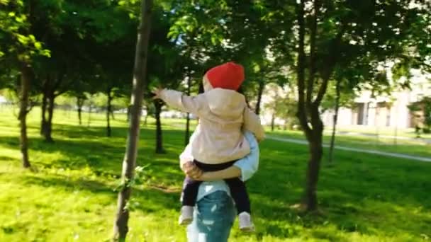 Ευτυχισμένη μητέρα κρατά το παιδί στην αγκαλιά της, σε ένα πάρκο της πόλης. Μωρό παίζει με ένα φύλλο στο δέντρο στο ηλιοβασίλεμα. — Αρχείο Βίντεο