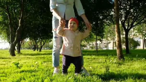 Ευτυχισμένο παιδί μαθαίνει να περπατάει στο πάρκο στο ηλιοβασίλεμα. Η μητέρα βοηθά το μωρό να λαμβάνει την πρώτα βήματα — Αρχείο Βίντεο
