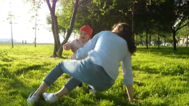 Matka i dziecko są gry w zachodzie słońca parken. Mama leży na trawie i podnosi stopy dziecka nad głową — Wideo stockowe