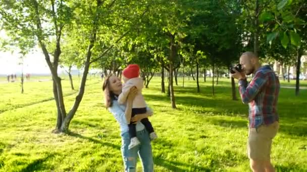 Jovem pai tirando fotos de mãe e filha brincando no parque ao pôr do sol. Família feliz tendo um resto e fotografado na natureza — Vídeo de Stock