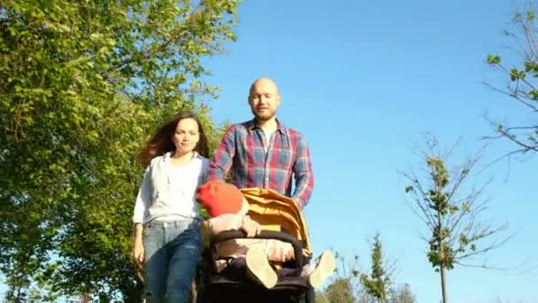 Pais caminhando com uma criança no parque. Família feliz: mamãe e papai rolaram criança no carrinho na natureza ao pôr do sol — Vídeo de Stock