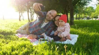 Mutlu aile üzerinde çim döşeme ve selfie bir bebekle günbatımında parkta yapıyor. Babam ve annem telefonda bebek ile kendilerini fotoğraf çekmek