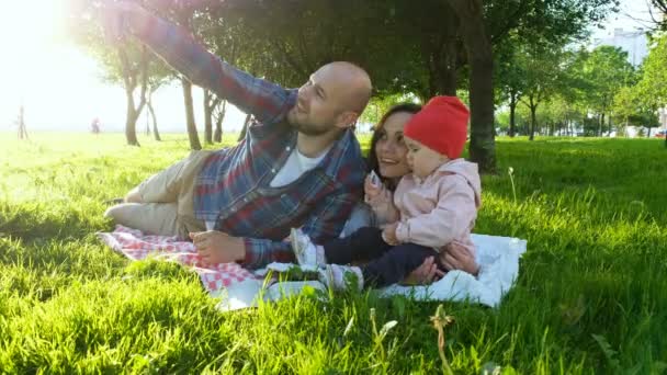 Šťastná rodina leží na trávě a otec dělá selfie s dítětem při západu slunce v parku — Stock video