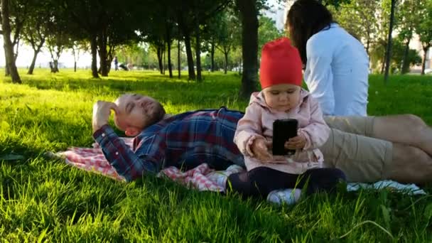 A menina está brincando com um smartphone na natureza. Família jovem está descansando no parque — Vídeo de Stock