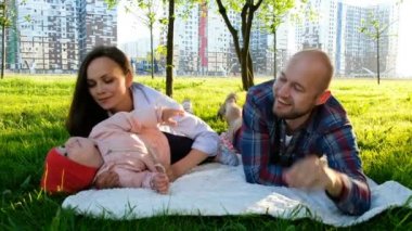 Genç Anne baba gülüyor ve saatler yaz aylarında parkta küçük kızı öper. Küçük bebek günbatımında yaz Park'ta dinlenme ile mutlu aile