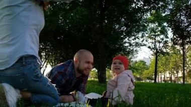 Yaz gün batımında parkta oturan genç bir çocuk ile mutlu bir aile. Çimenlerin üzerinde otururken gülüyor küçük bebek kız