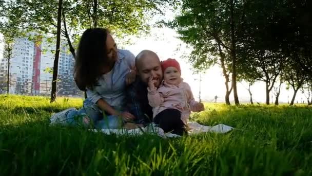 Família feliz brincando com criança no parque de verão ao pôr-do-sol. Uma menina pequena ajusta o nariz de seus pais e ri — Vídeo de Stock