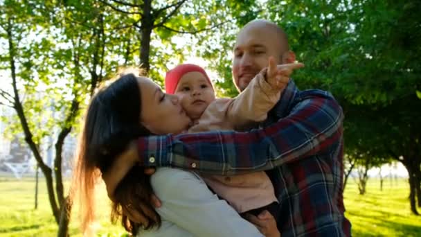 Una familia joven con un niño pequeño se abraza y se besa. Padres de familia sostienen a su hija en brazos en el parque en el verano al atardecer . — Vídeos de Stock