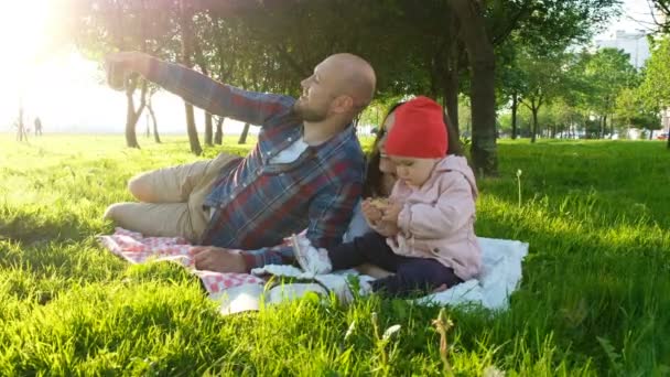 La familia feliz está acostada en la hierba y el padre haciendo selfie con un bebé al atardecer en el parque en el teléfono inteligente — Vídeo de stock