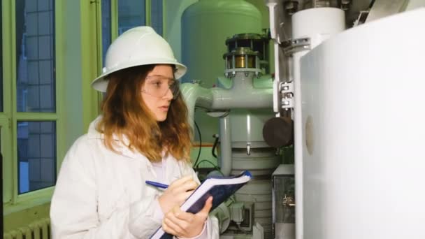En flicka ingenjör i en vit hjälm skriver ner en danne med en kemisk industrianläggning i ett laboratorium för produktion. — Stockvideo