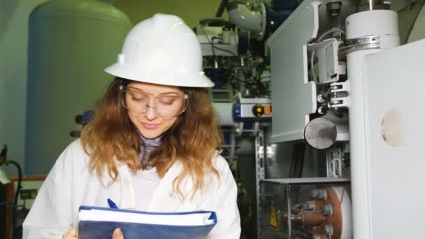 Bella ragazza scienziata che fa un diario in un laboratorio di produzione, su uno sfondo di apparecchiature a gas, guardando la fotocamera e sorridendo . — Video Stock