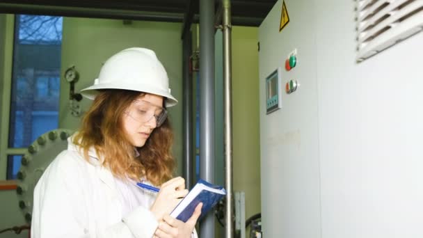 Uma engenheira em um capacete branco usando um vestido e óculos de segurança registra os dados da exibição do armário de controle elétrico na sala de produção . — Vídeo de Stock