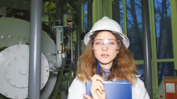 Menina engenheiro em óculos e um capacete branco pensa e olha para a câmera, segura uma caneta e pasta com papéis, contra o fundo do equipamento de gás . — Vídeo de Stock