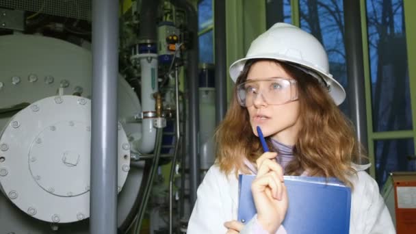 Une belle fille ingénieur dans un casque blanc pense sur le fond de l'équipement à gaz, elle a eu l'idée et elle l'a écrit dans un magazine . — Video