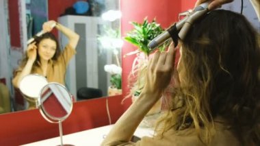 Genç güzel kız aynaya bakar ve bukle maşası soyunma odasında yumuşak odak üzerinde bukleler